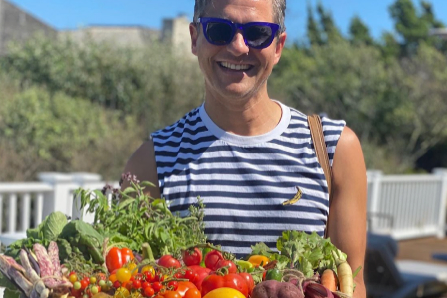 David Burtka holding a basket of produce, wearing a blue and white striped shirt and sunglasses, smiling