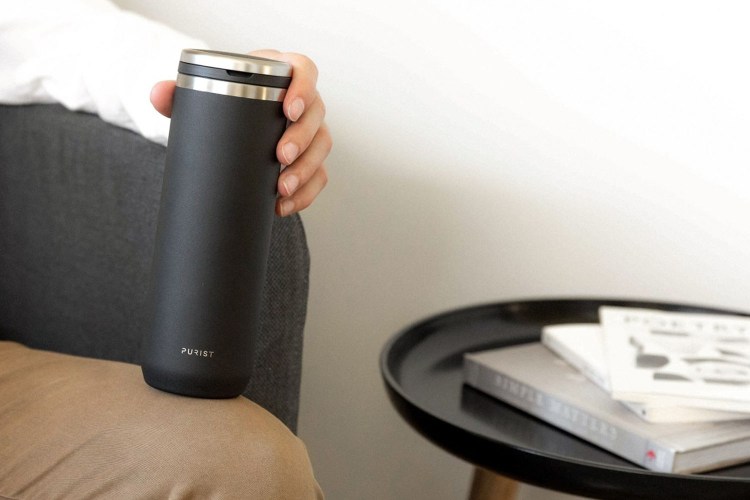 Hand holding a black Purist Mover water bottle while sitting next to a side table with books