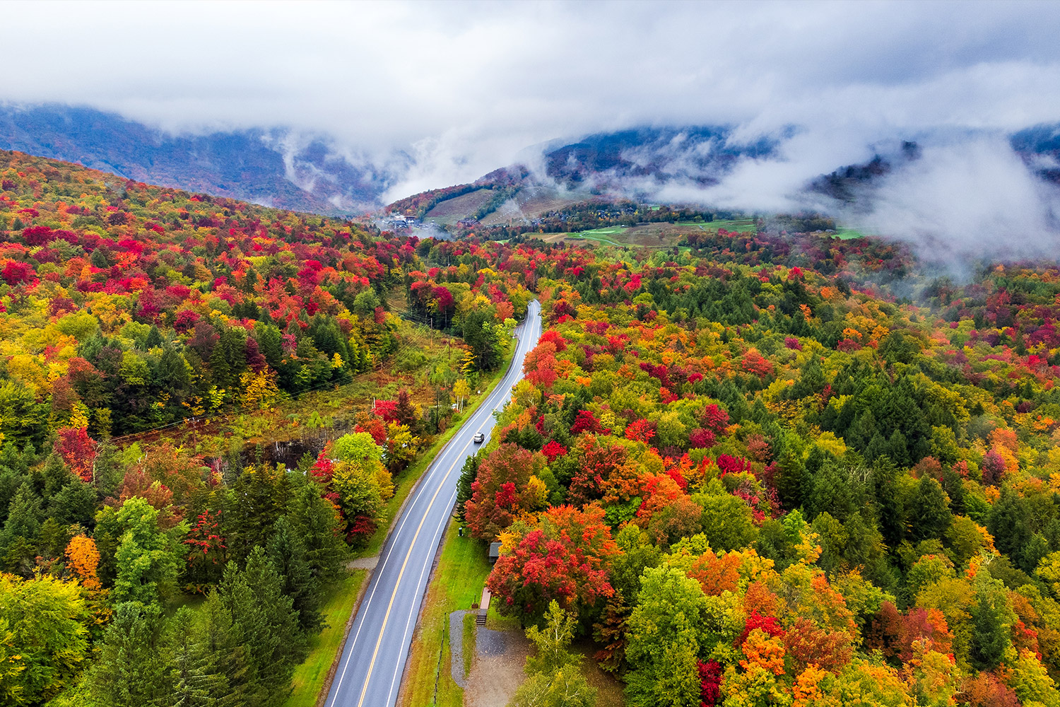 View from above road showing leaves starting to change deep red, orange, and yellow. Hills covered by clouds in back