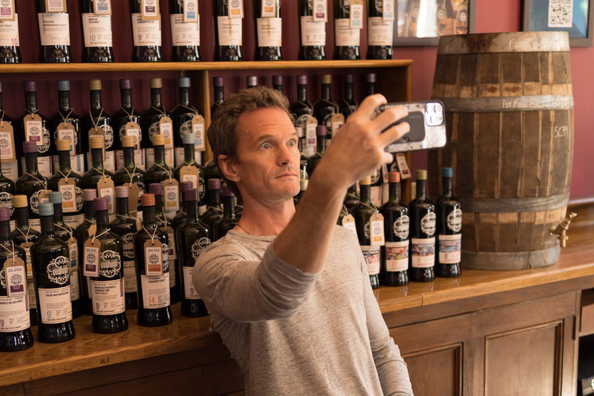 Neil Patrick Harris with eyes wide taking a selfie in front of a wall full of liquor