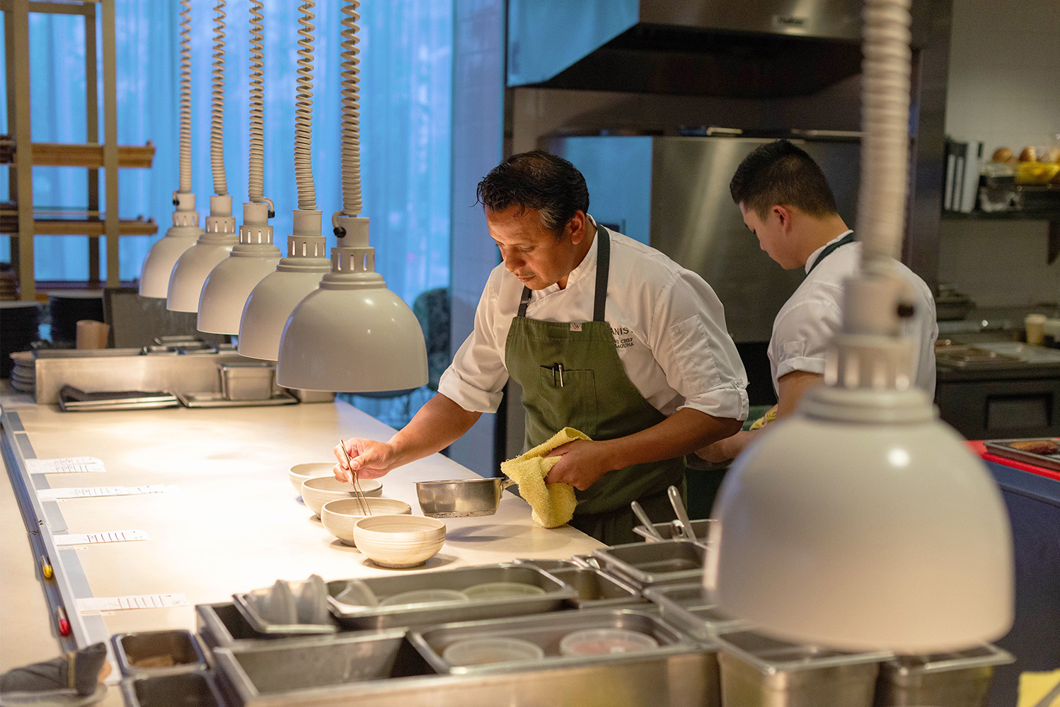 Chef Hector Laguna in chef jacket and apron plating food with chopsticks in an industrial kitchen