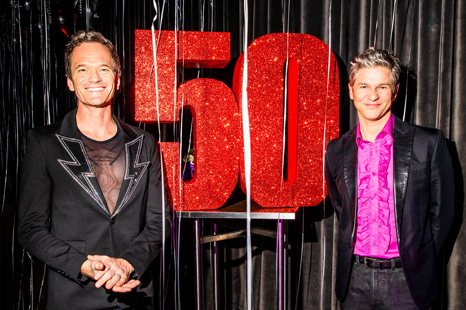 Neil Patrick Harris and husband, David, smiling next to giant, sparkly red 50