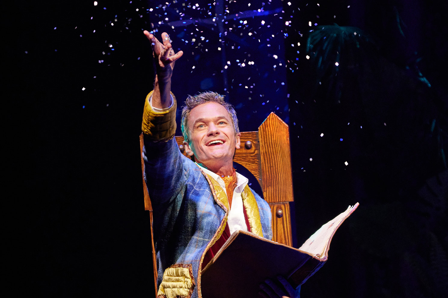 Neil sitting in wooden chair in costume on stage with arm raised as glitter falls from sky