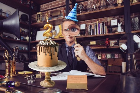 Neil Patrick Harris in office holding a magnifying glass while wearing a party hat. Chocolate cake with "2" candle is on the desk