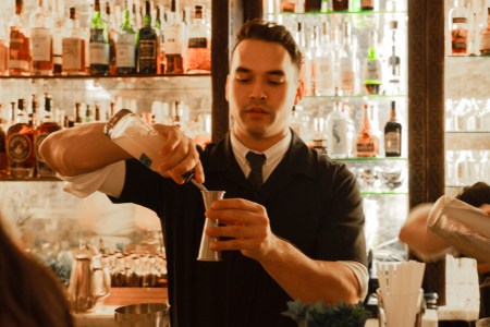 Mixologist and bartender, Jonathan Lind, pouring flavored syrup into a jigger in front of a lit up wall full of liquor bottles