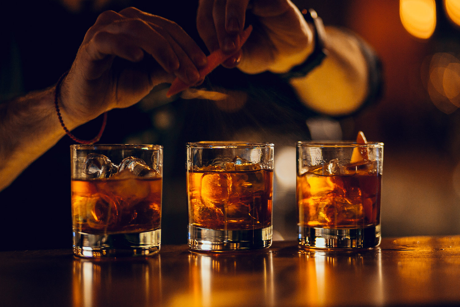 A bartender expressing an orange peel over three finished old-fashioned cocktails