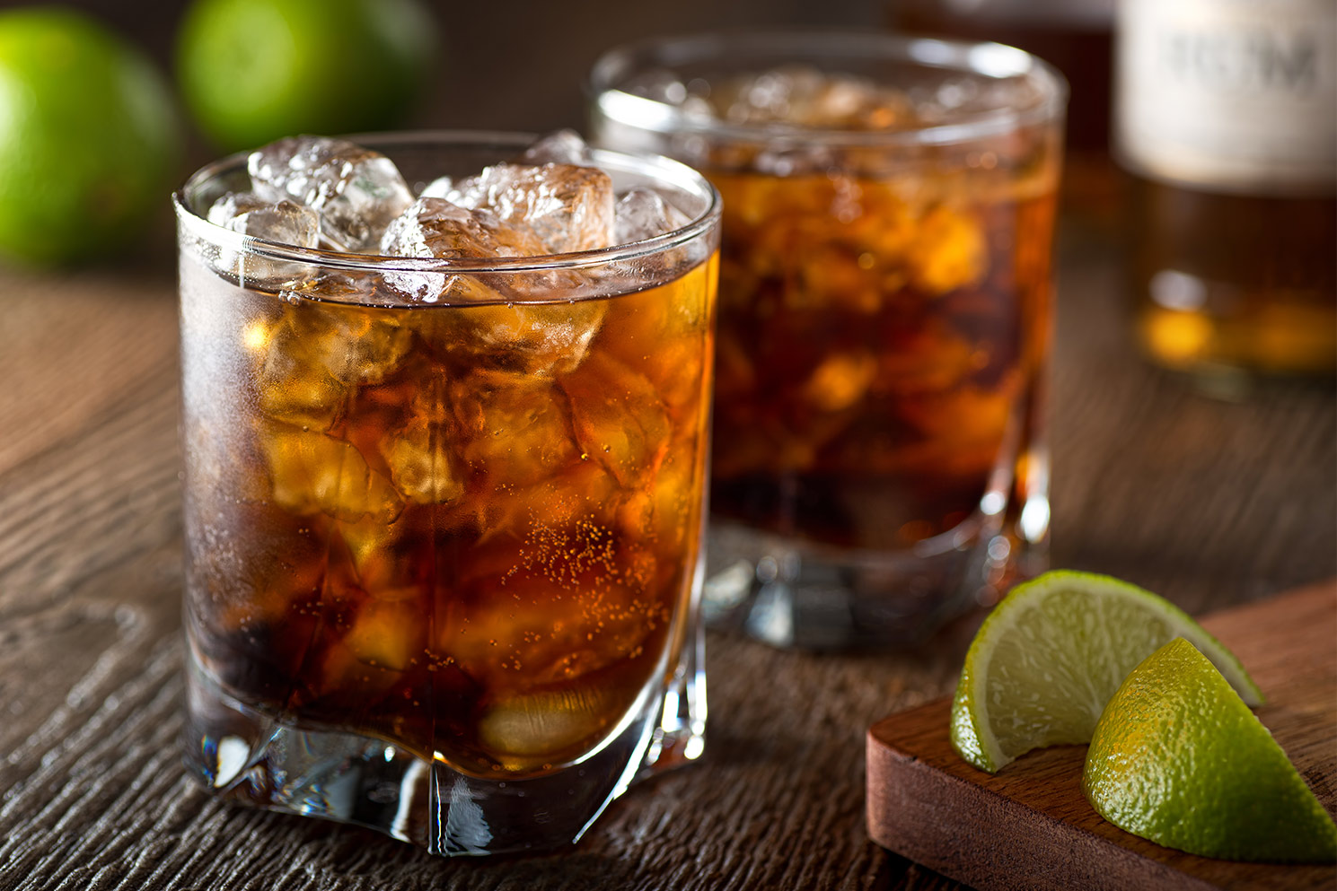 Two rum and cokes full of ice on wood table with lime wedges