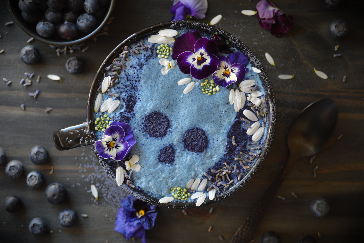 A blue latte with skull art in the milk foam decorated beautifully with flowers and petals. Shown from above with blueberries surrounding latte