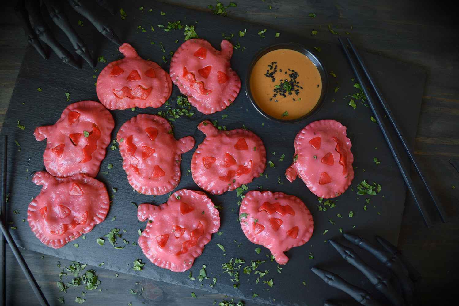 Pink pumpkin shaped potstickers with red jack-o-lantern faces on a matte black board with an orange sauce and black chopsticks