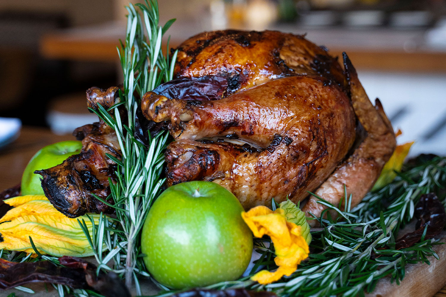 A glossy, charred turkey lays with rosemary garnish, yellow flowers, and green apples