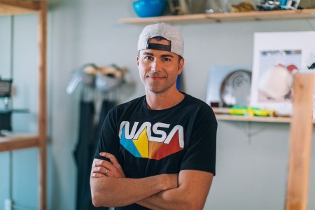 Mark Rober stands with arms crossed for a portrait wearing a backwards cap and a NASA graphic tee