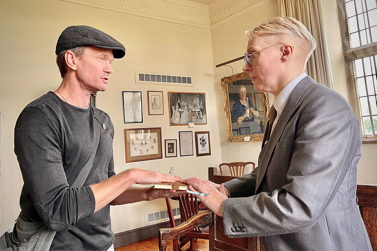 Neil Patrick Harris stands across from a person while they both have their hands on a book or journal