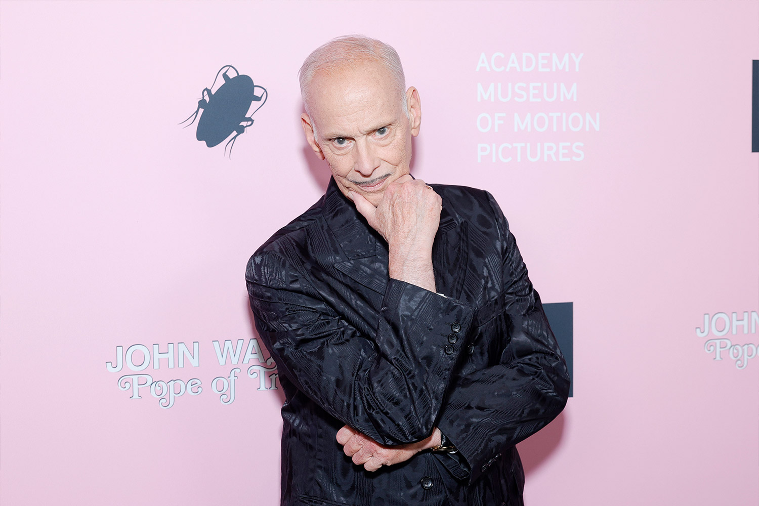John Waters holding his chin and posing in front of a pink background at an Academy Museum of Motion Pictures event