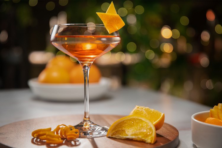An orange cocktail in a coupe glass with orange peel garnish